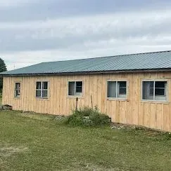 A long, single-story wooden building with light-colored vertical siding, a green metal roof, and several sliding windows.