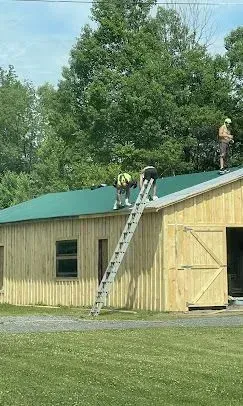 Three people working on the green metal roof of a light-colored wooden barn, with a ladder leaning against the side.