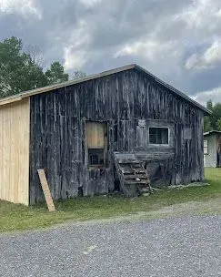 An old, weathered wooden shed with dark, vertical siding, a window, and a section of new, light-colored plywood paneling.