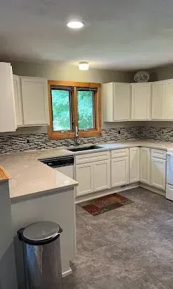 A kitchen with white cabinets, light countertops, a patterned tile backsplash, a window above the sink, and gray flooring.