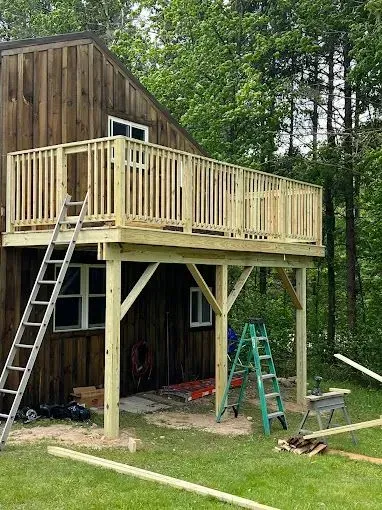 A newly constructed wooden deck attached to the second story of a brown, vertical-sided house amidst trees.