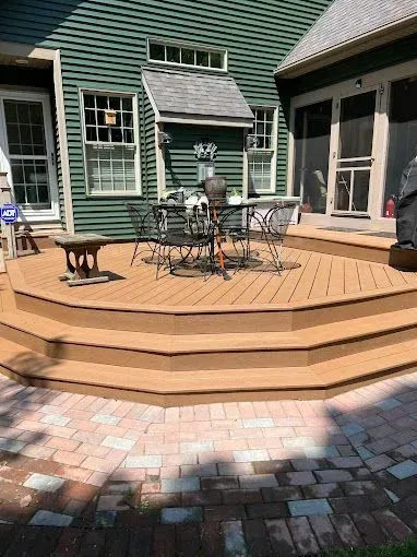 A tan-colored wooden deck with tiered stairs leads to a house with green siding and a patio made of red bricks.