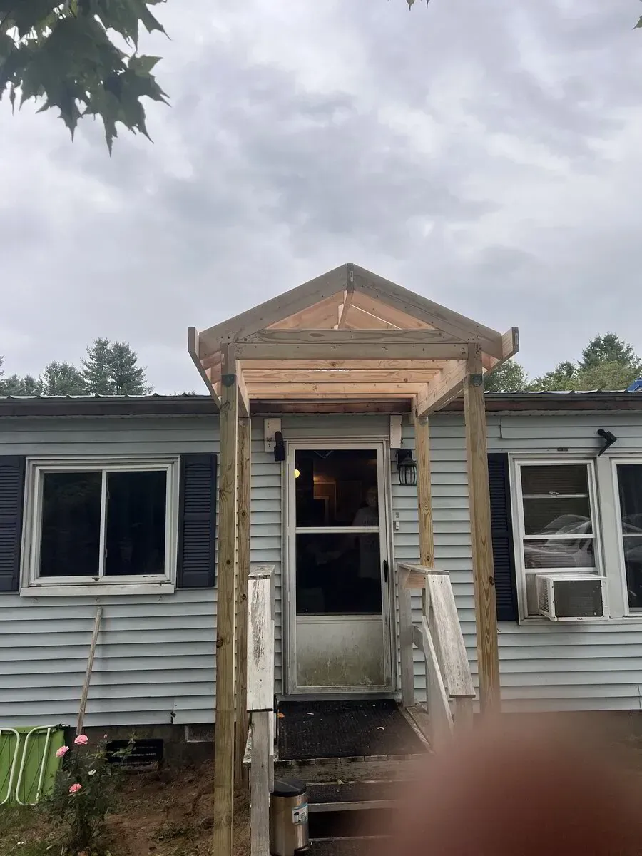 A wooden porch frame is under construction over the front door of a light blue mobile home with two side windows.