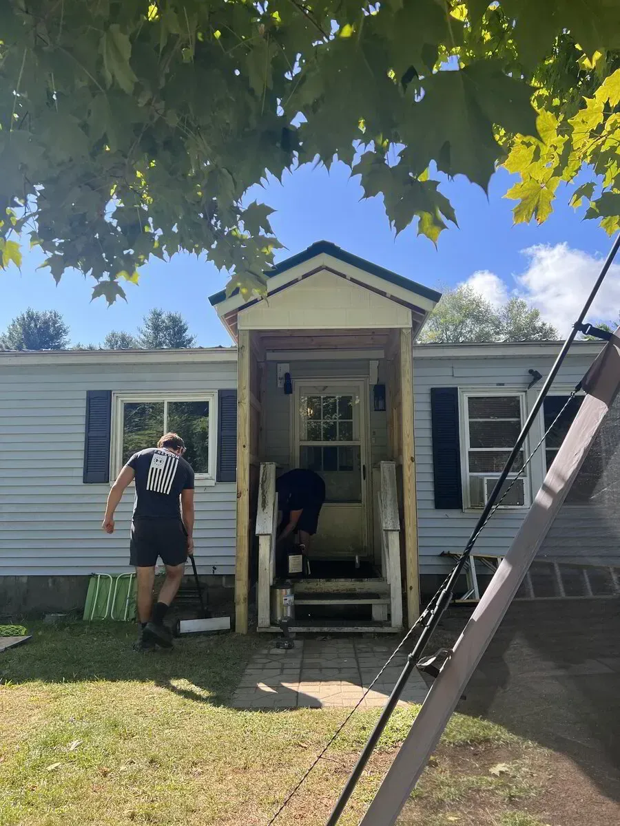 Two people work on a house exterior; one paints the siding while the other works inside the small, unfinished entryway.
