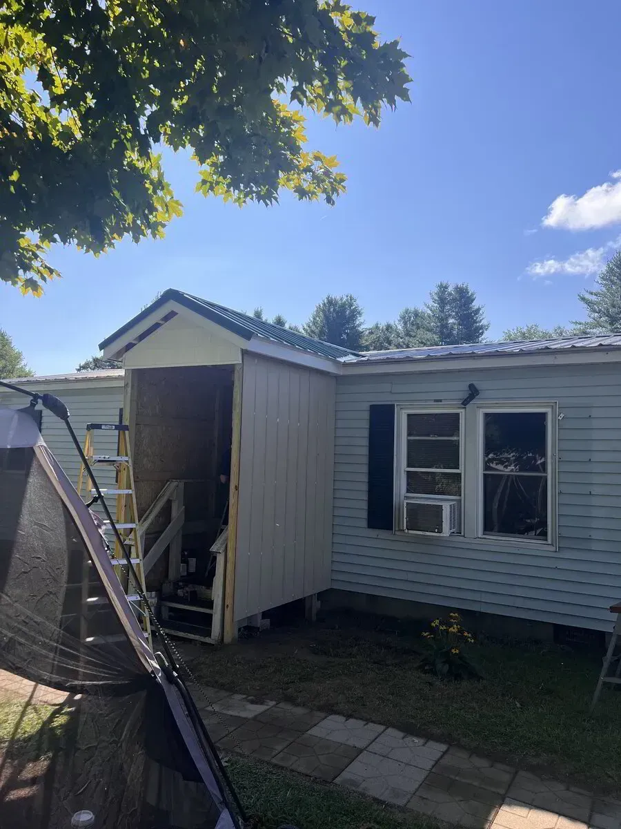 A light-colored mobile home with an unfinished entryway under construction, featuring a ladder and visible wooden framing.