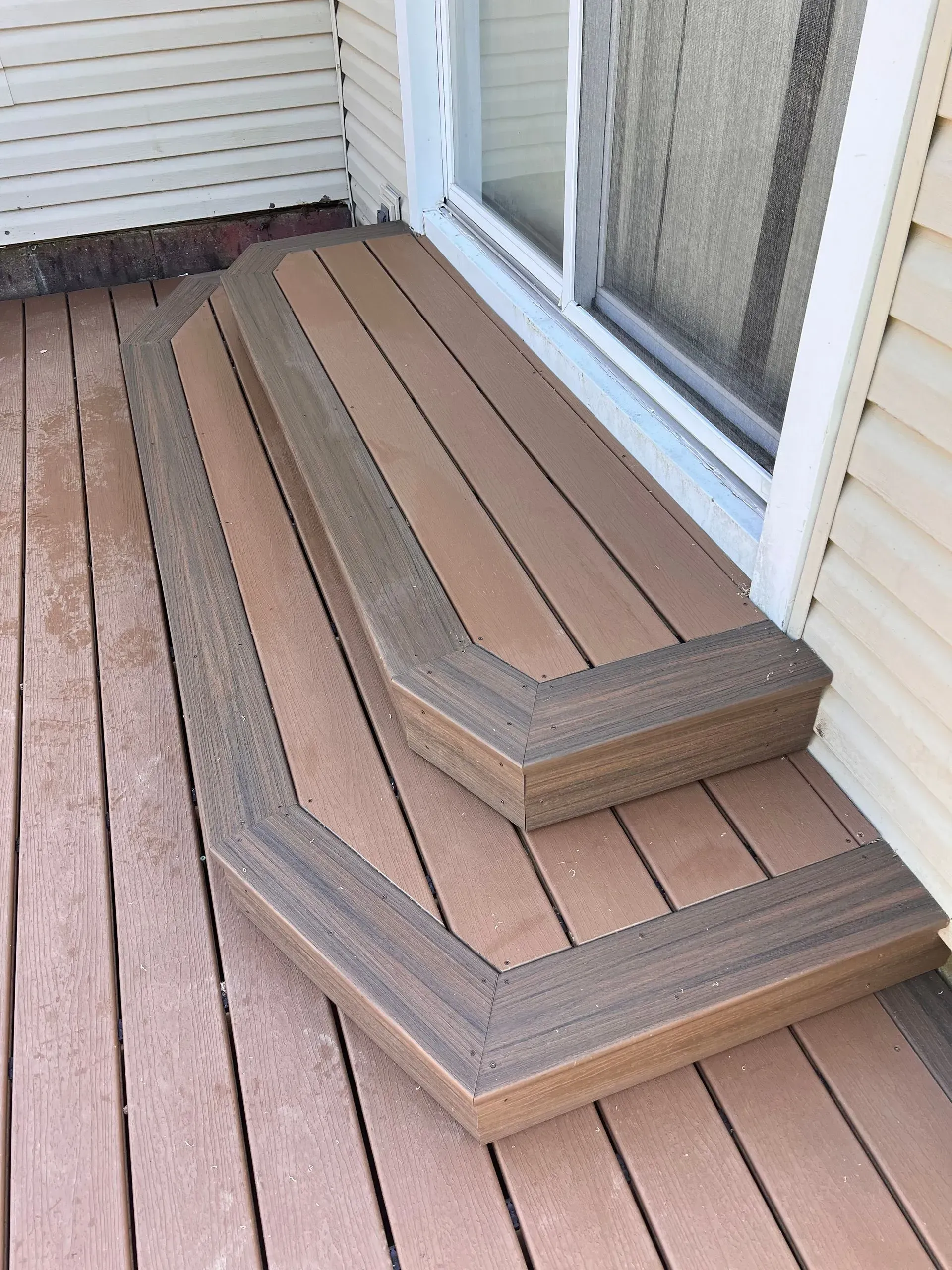 Two-step wooden porch stairs with light brown treads and dark brown trim leading to a sliding glass door on a deck.