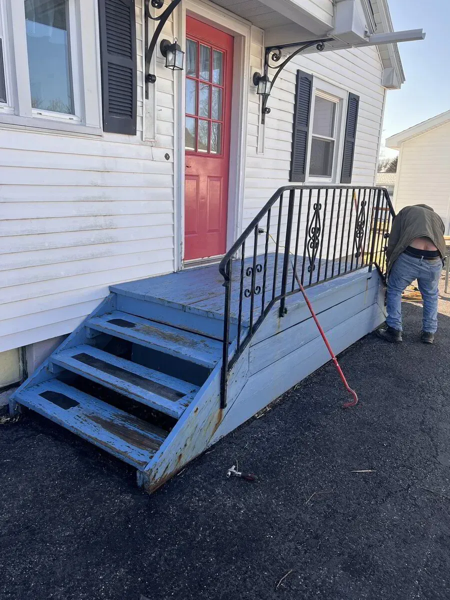 A person in jeans bends over to work on the side of blue wooden porch stairs in front of a house with a red door.
