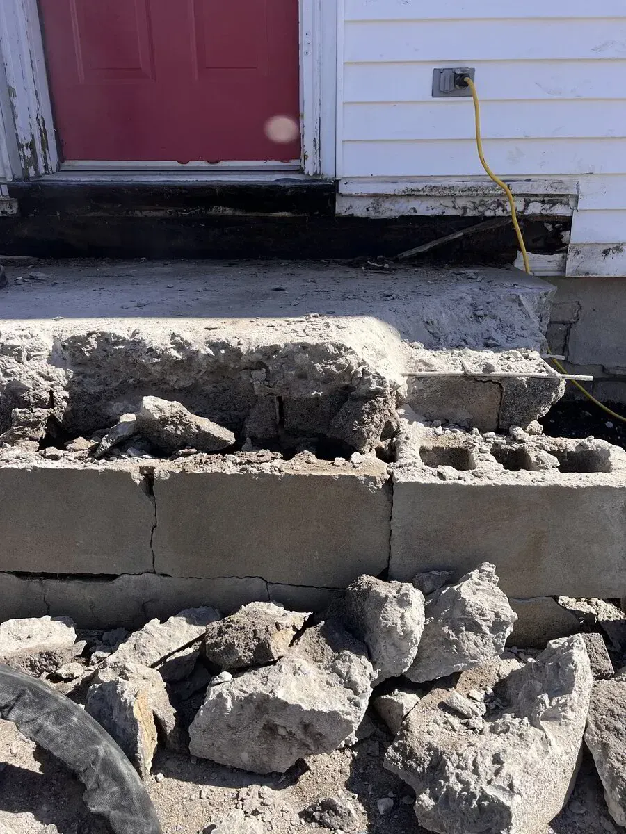 A crumbled concrete front porch step with exposed cinder blocks and scattered debris, leading to a red door.