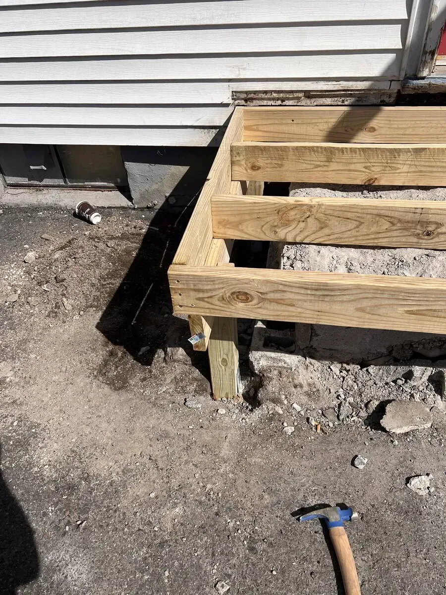 A wooden deck frame under construction against the side of a house, with a hammer resting on the ground nearby.