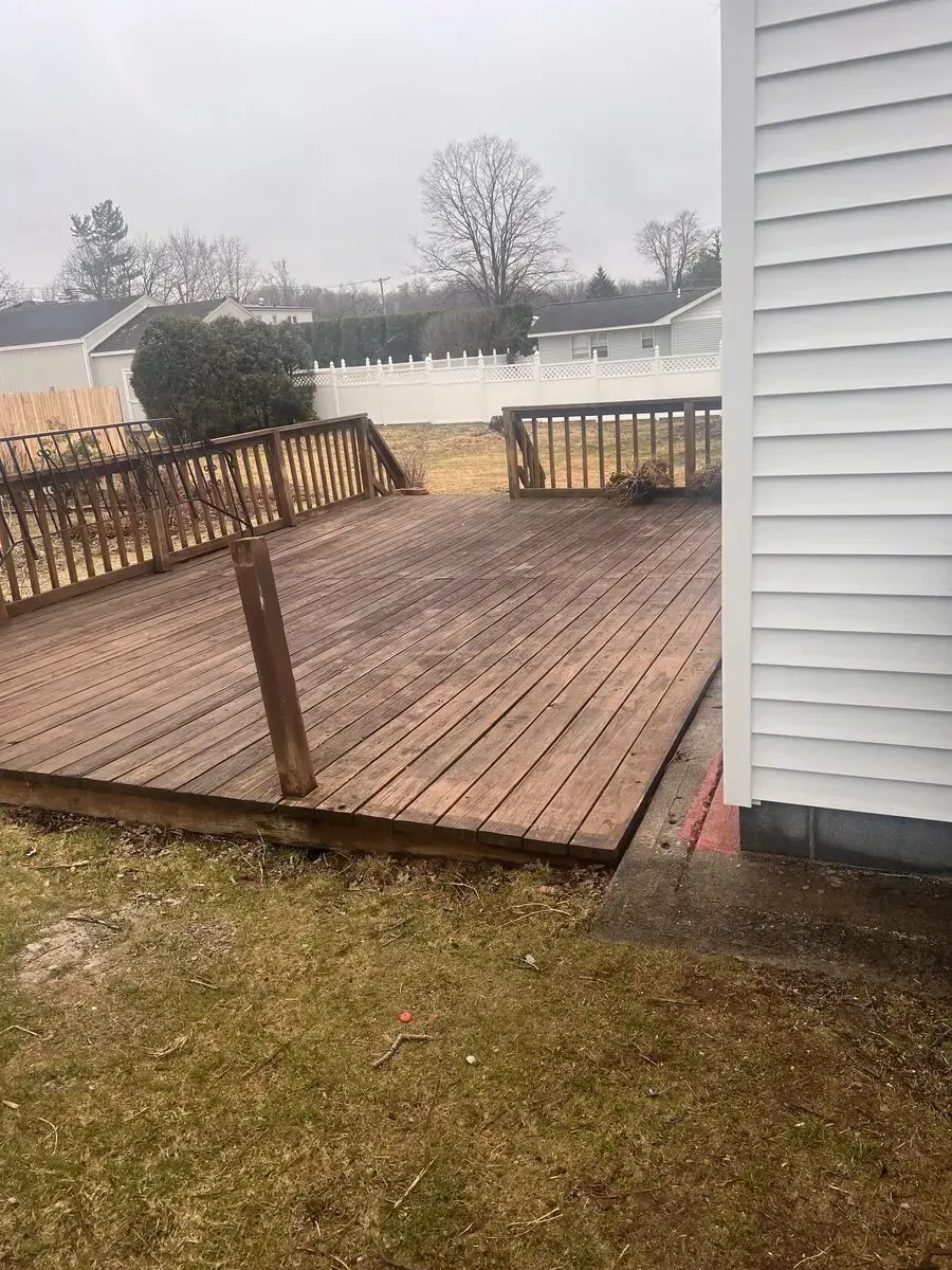 A wooden deck attached to a house, with railings and a white fence in the background on a cloudy day.