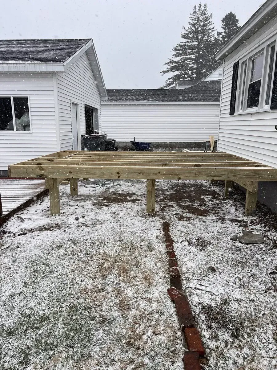 A wooden deck frame under construction in a snowy backyard between two white houses.