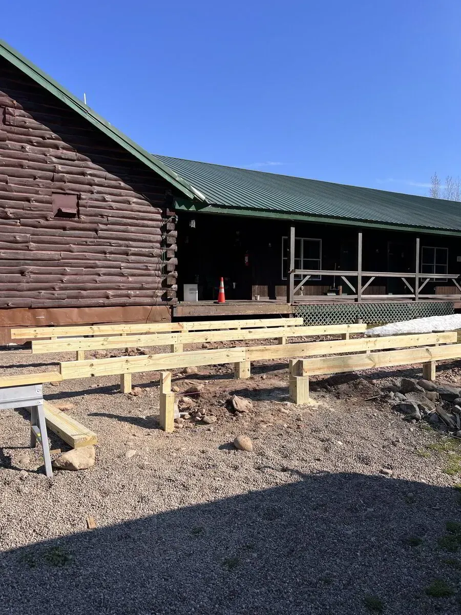 A brown log building with a green metal roof under construction with wooden framing for a deck on a gravel ground.
