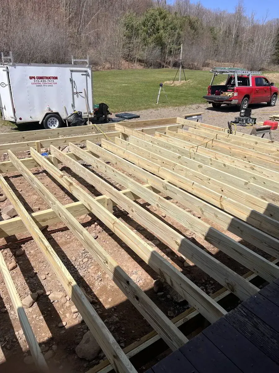 Wooden deck joists installed over a dirt ground, with a utility trailer and a red truck parked in the background.