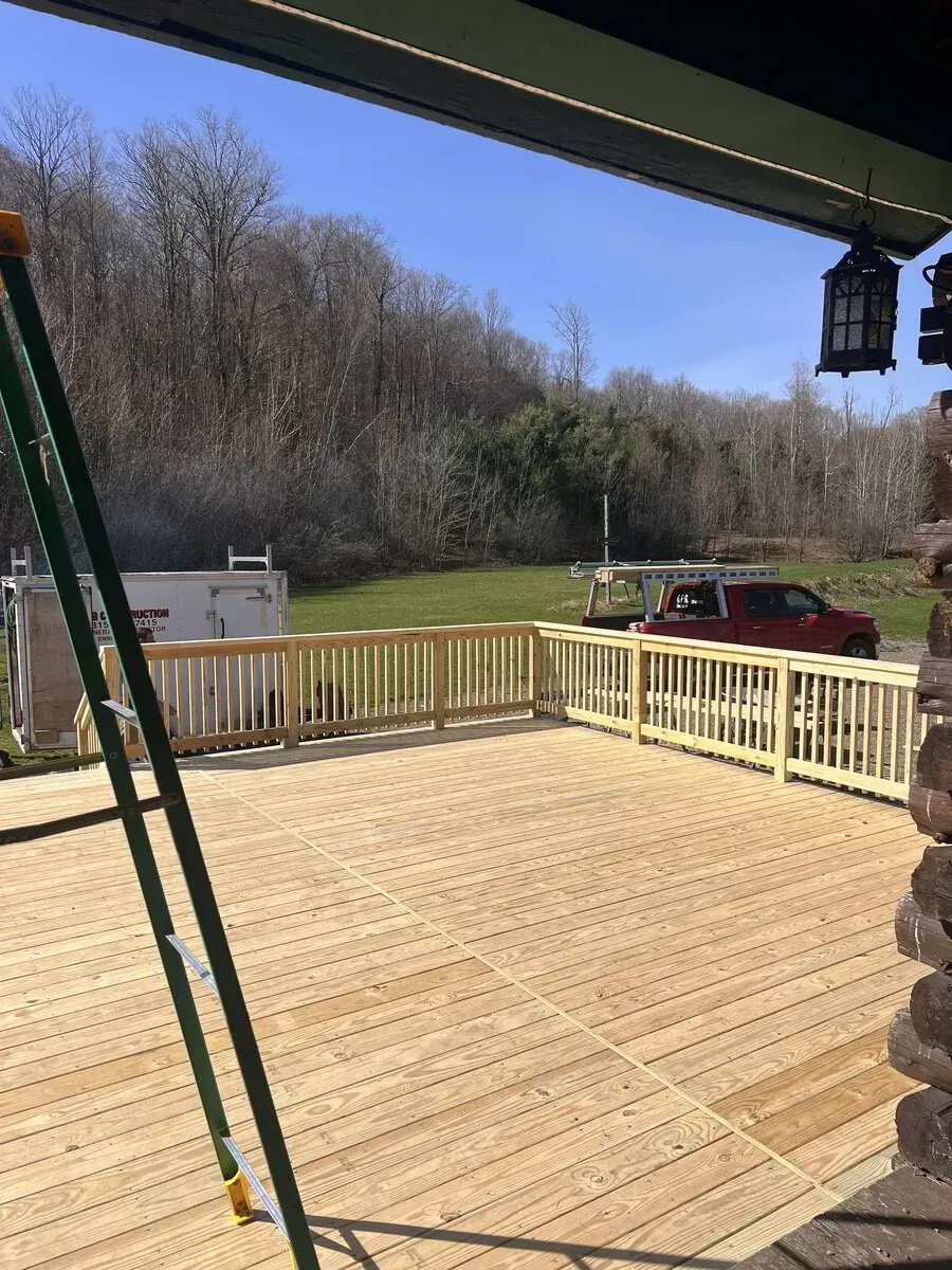 A newly constructed wooden deck with railings, viewed from a porch, overlooking a field with trees and a red truck.