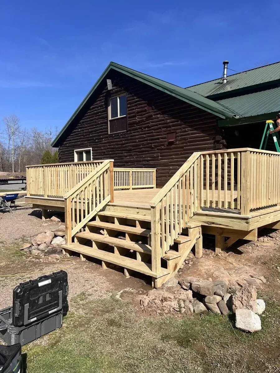 A freshly built wooden deck with stairs attached to a dark-sided cabin under a clear blue sky.