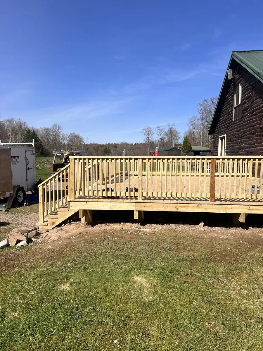 A newly constructed wooden deck with railings attached to the side of a brown house on a sunny day.