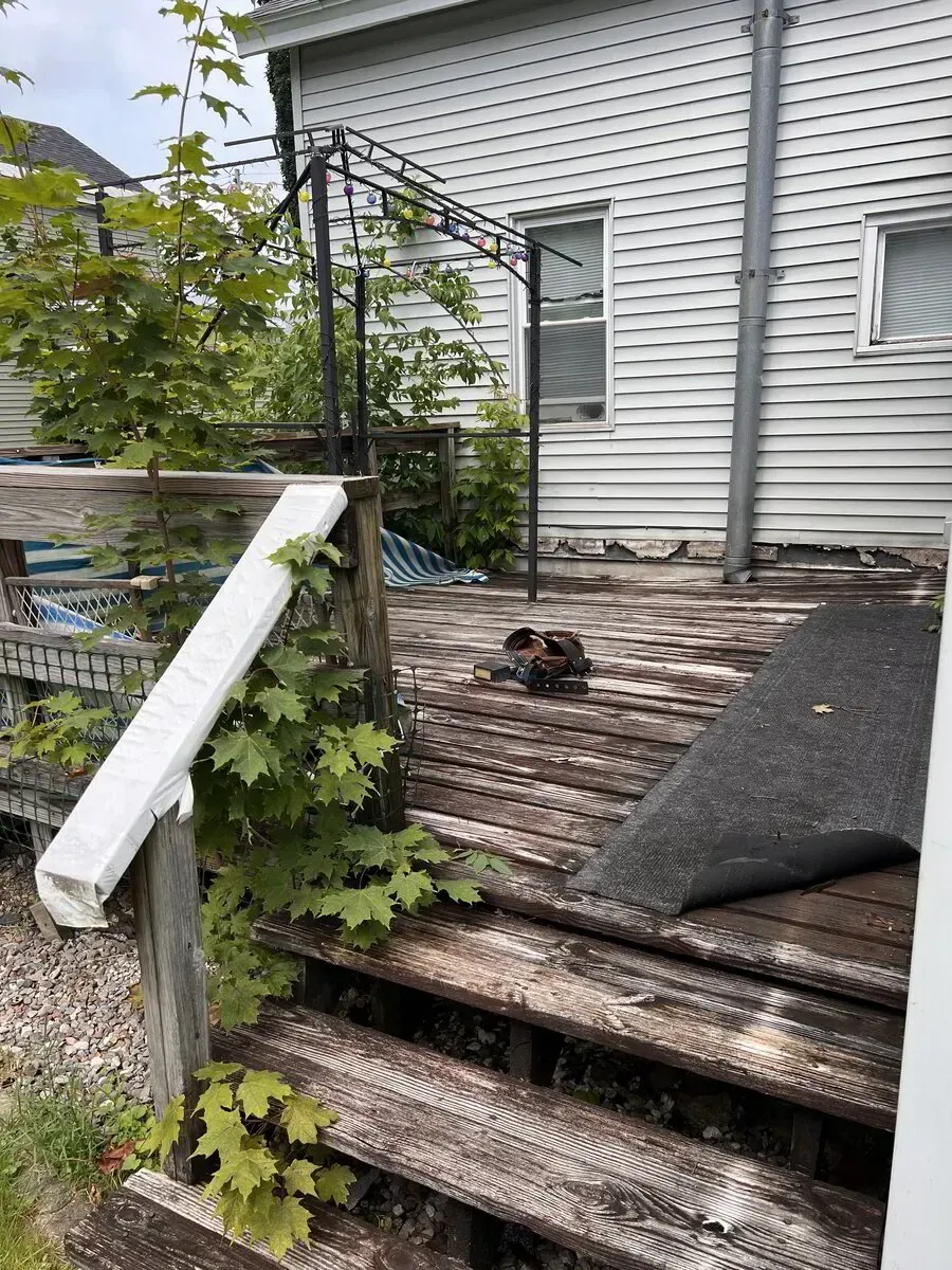 A weathered wooden deck and stairs outside a house, with an overgrown vine on the railing and a dark mat on the floor.