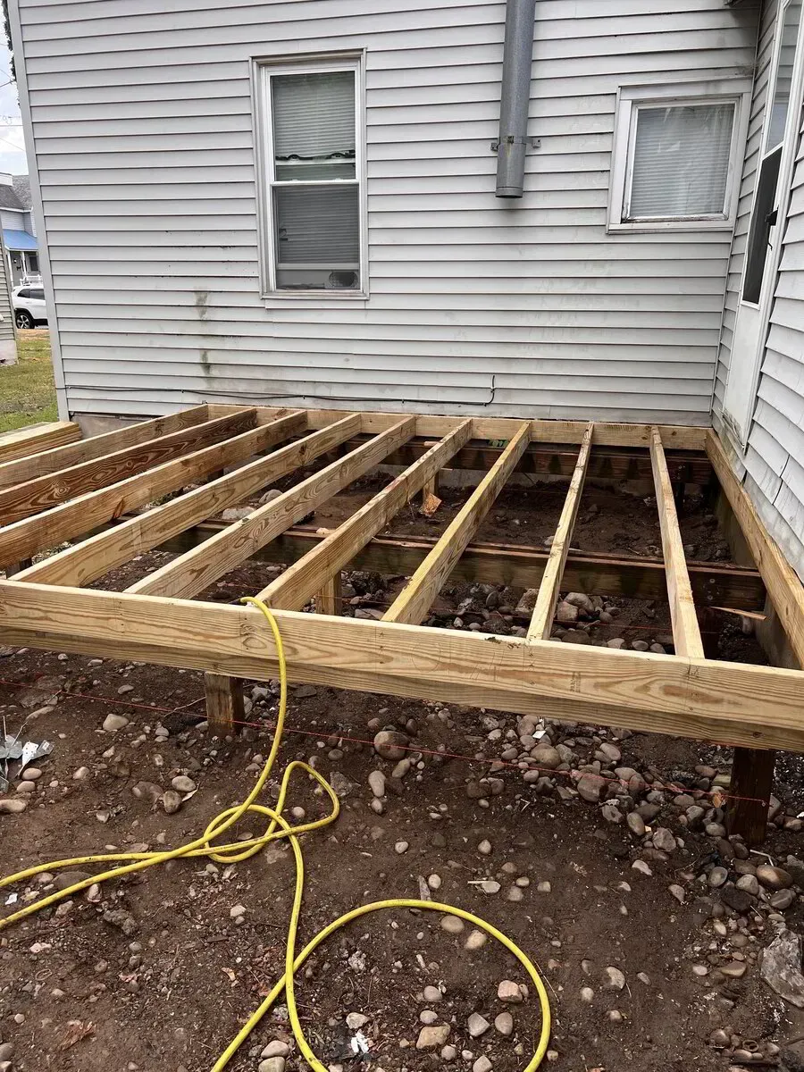 A wooden deck frame is under construction against the side of a house with grey siding, over a dirt and gravel ground.