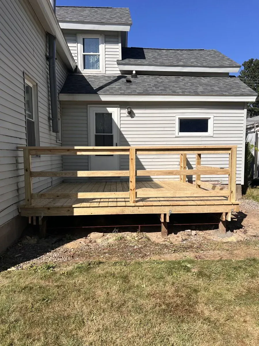 A newly constructed, light-colored wood deck with railings sits against the light gray siding of a two-story house.
