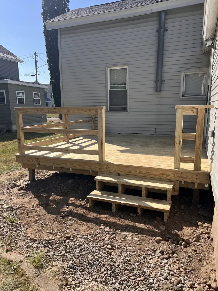 A new, light-colored wooden deck with railings and steps attached to the back of a grey, vinyl-sided residential home.