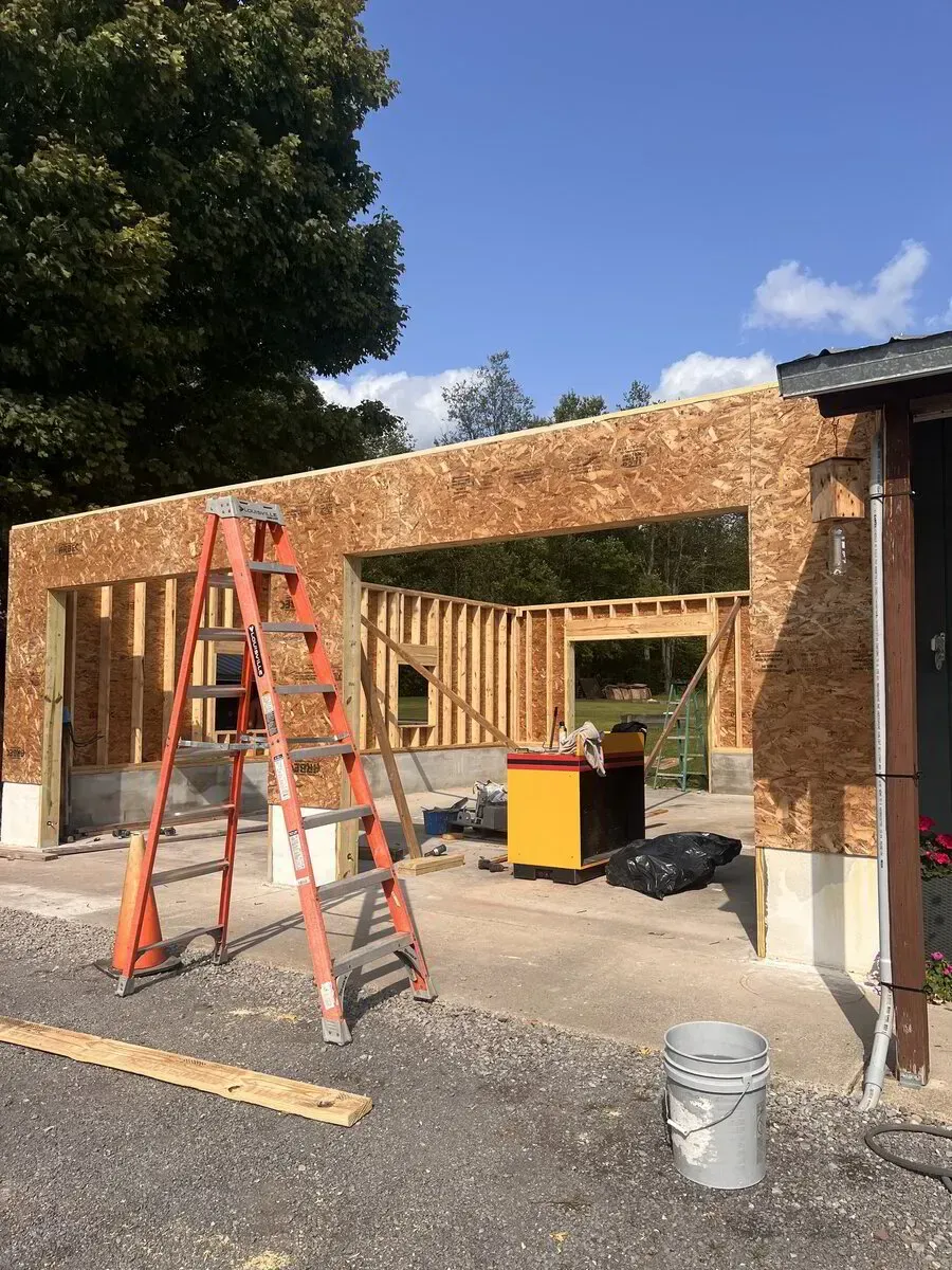 An orange ladder stands in front of a building under construction with exposed wooden framing and a large open doorway.