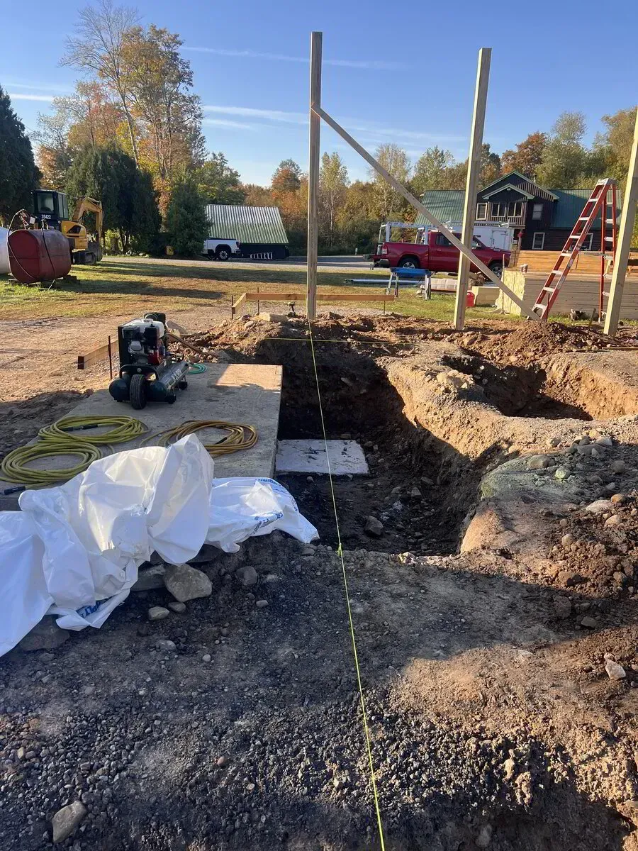 An outdoor construction site shows a deep excavated trench, two vertical wooden posts, and a ladder against a frame.