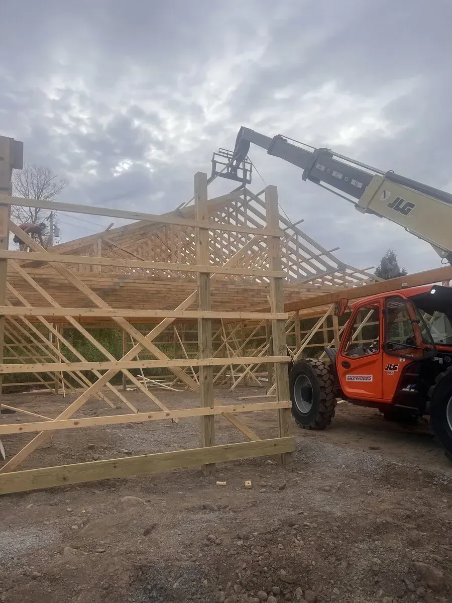 An orange telehandler lifts a worker in a platform to the roof frame of an unfinished wooden building on a cloudy day.