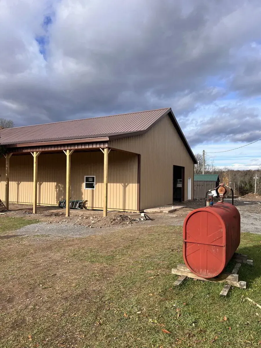 A tan pole barn with a metal roof and open porch, with a red metal tank sitting in the foreground on a grassy lot.