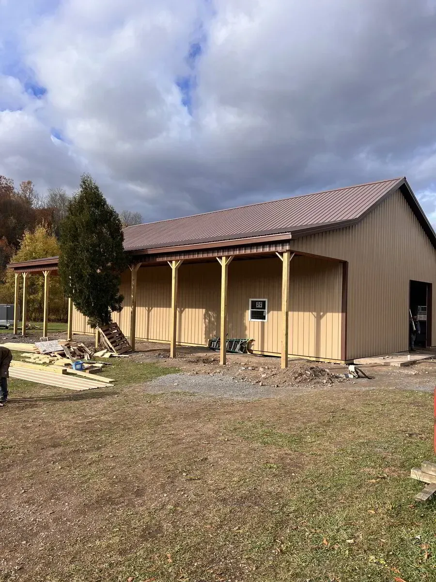 A tan metal pole barn with a brown roof and an open porch sits on a gravel lot under a cloudy sky.