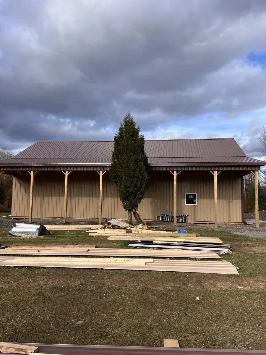 A newly constructed tan metal building with a dark roof and wooden posts, featuring a tall evergreen tree in front.