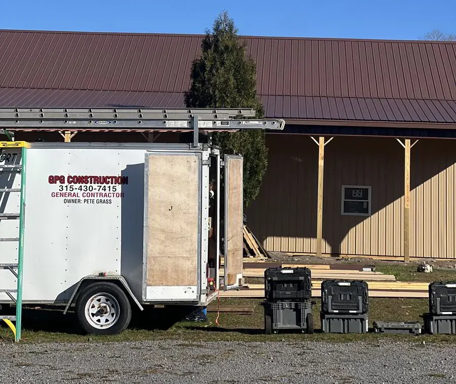 A white GPS Construction utility trailer sits on a gravel lot next to a building with a brown roof and tan siding.