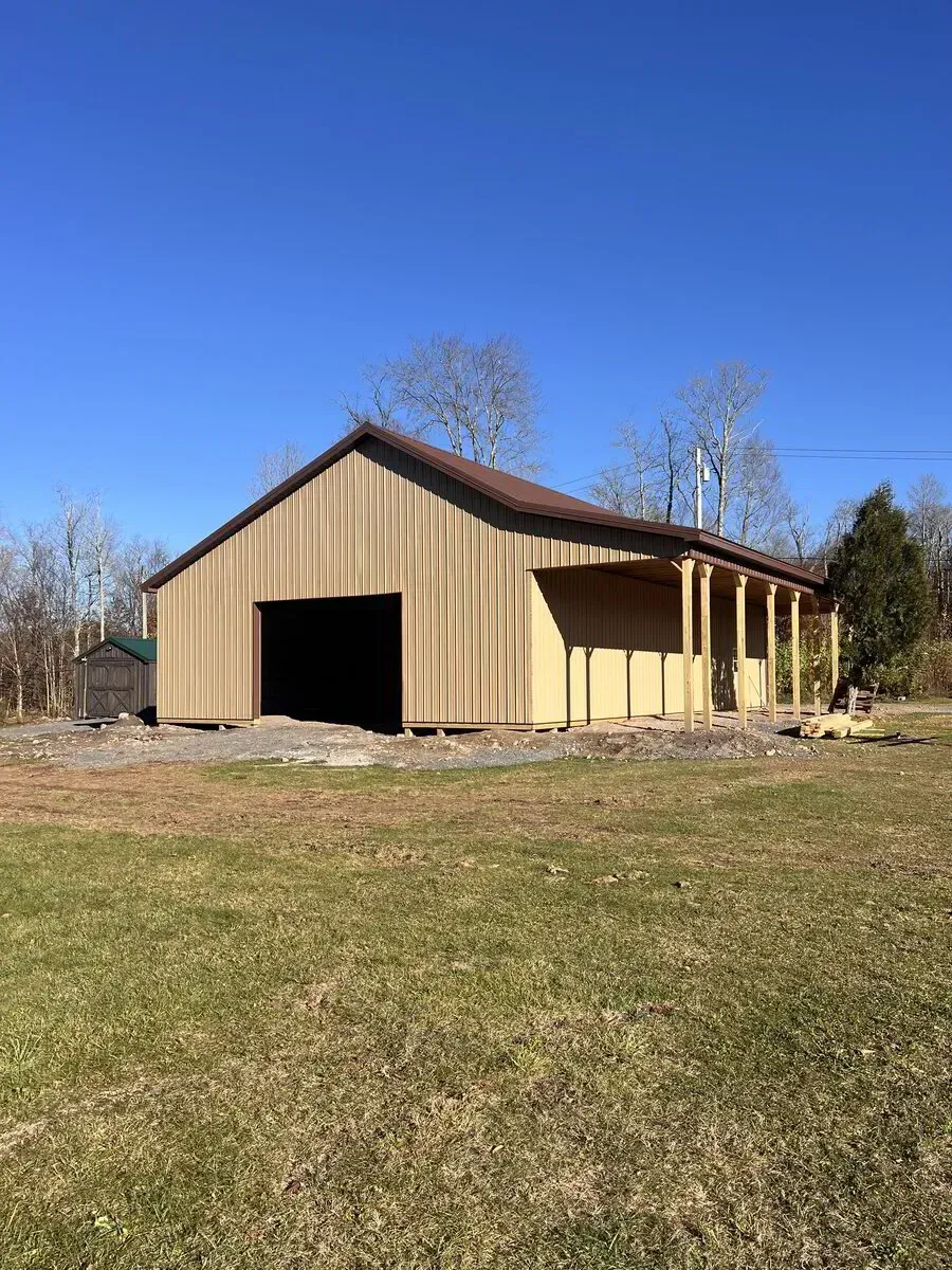 A beige barn with a brown roof and an open garage bay sits on a grassy field under a clear blue sky.