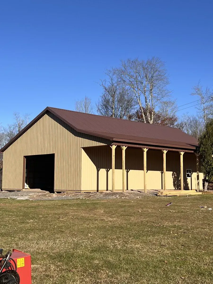 A tan metal barn with a brown roof and an open-air side porch stands in a grassy field under a clear blue sky.