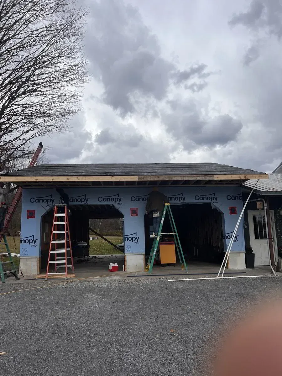 Workers renovate a garage exterior with light blue sheathing, using ladders under a cloudy sky.