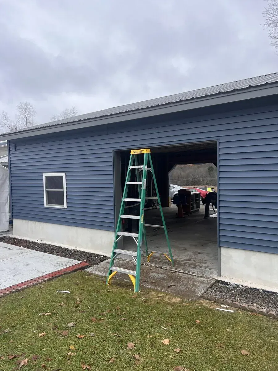 A green stepladder stands in front of the open bay of a blue-sided garage on a grassy lot under an overcast sky.