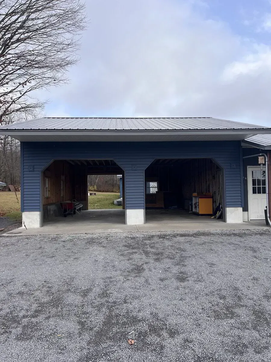 A blue two-bay open garage with a metal roof, white concrete pillars, and an adjacent white door on a gravel driveway.