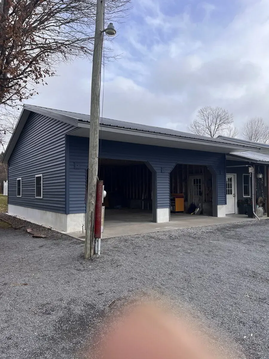 A blue garage with a white foundation and two open bays, standing on a gravel driveway under a cloudy sky.