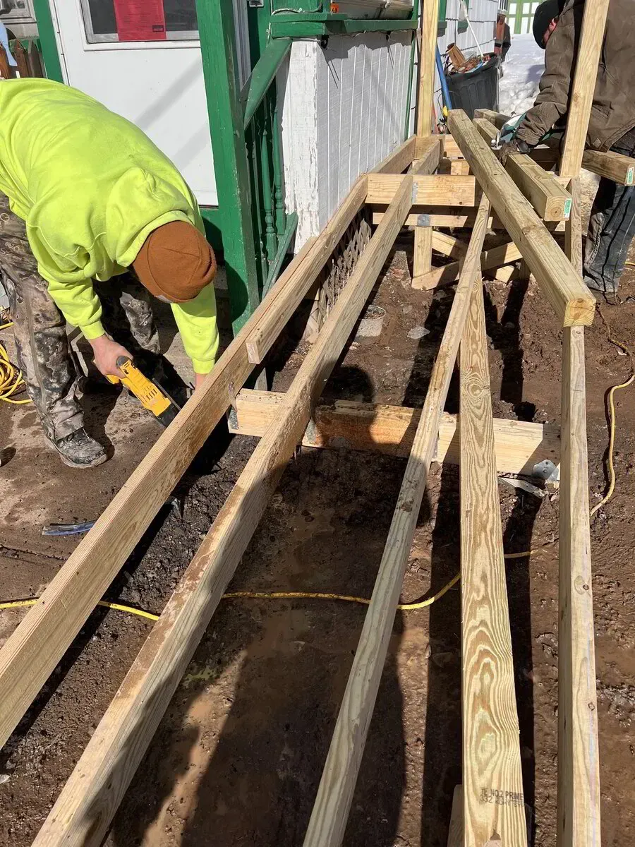 A person in a bright yellow hoodie uses a power drill on a wooden deck frame under construction outdoors.