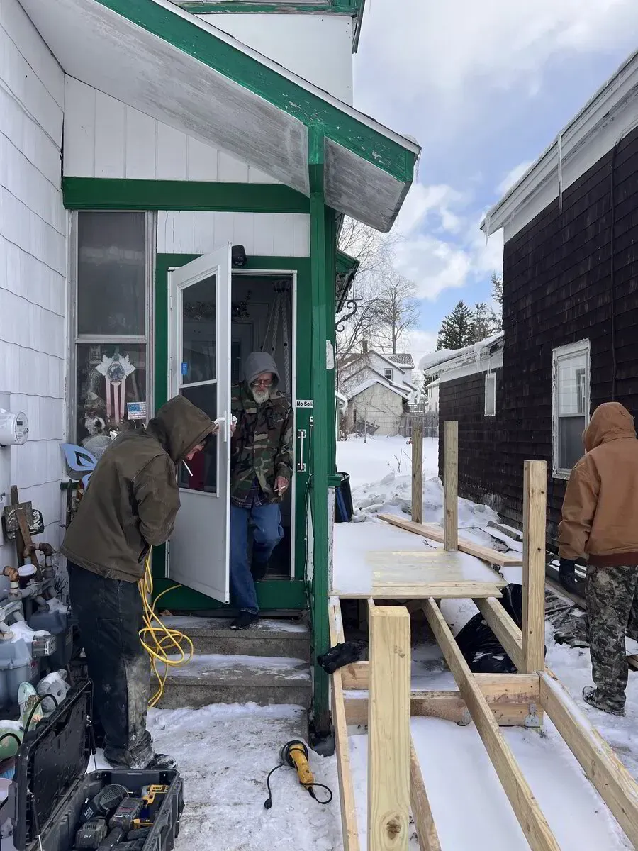 Three people wearing winter gear work on constructing a wooden accessibility ramp outside a snow-covered home.