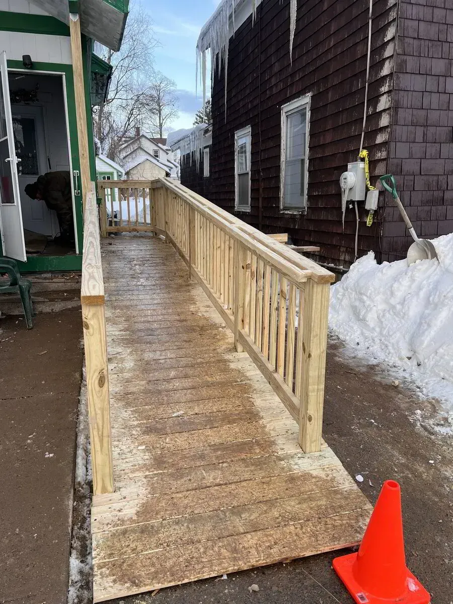 A newly built wooden wheelchair ramp leading up to a house entrance, with an orange safety cone on the snowy ground.