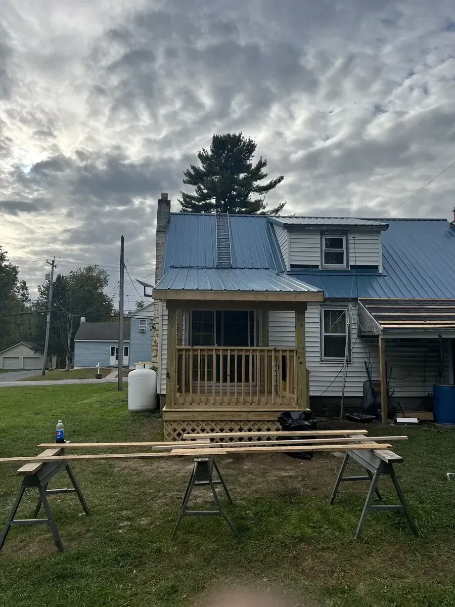 A house with a newly built wooden porch and metal roof, under construction in a yard with sawhorses in the foreground.