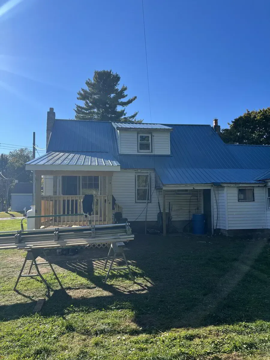 A white house with a bright blue metal roof, featuring a partially built wooden porch and a dormer window under a blue sky.