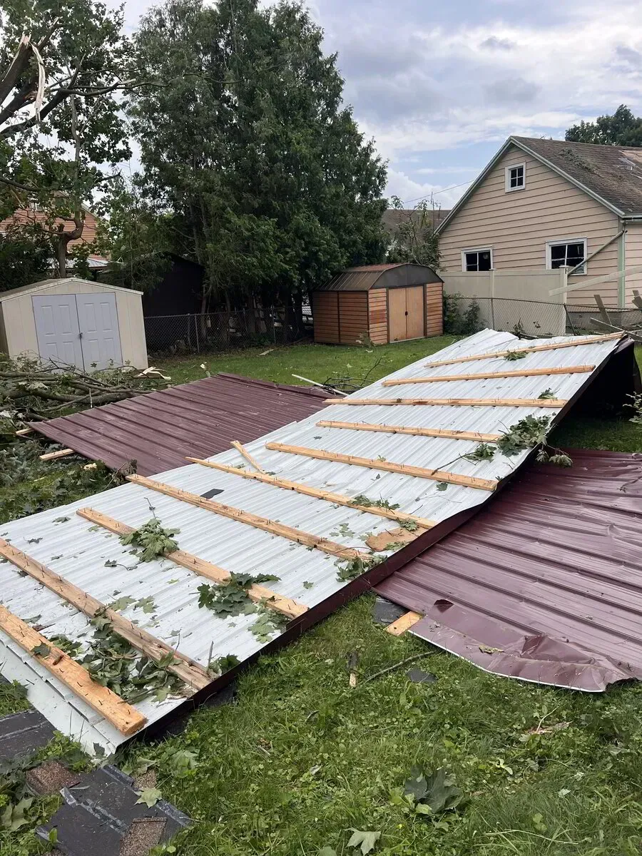 A large metal roof panel lies collapsed on a grassy backyard near storage sheds and a house after a storm.
