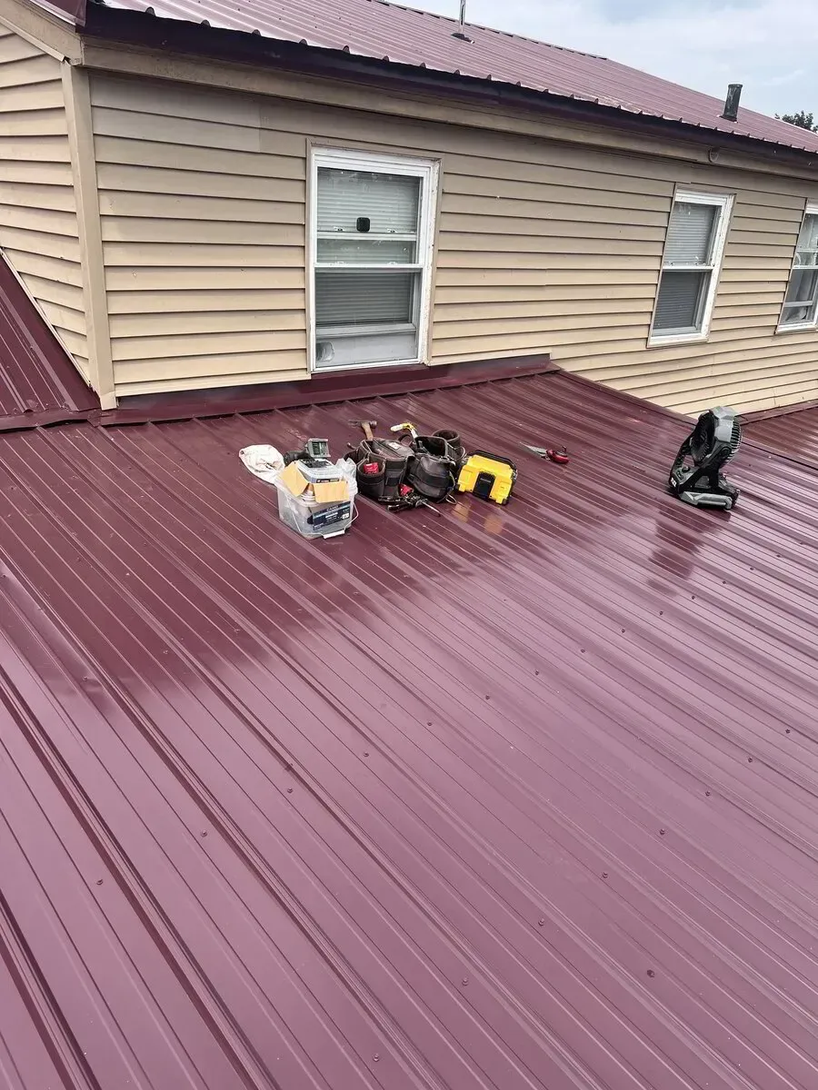 Tools and a white bucket sit on a red corrugated metal roof below the beige siding and windows of a house.