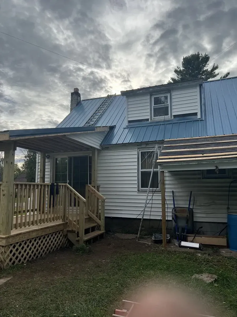 A blue metal-roofed house under a cloudy sky, featuring a newly built wooden deck and partial exterior siding repairs.