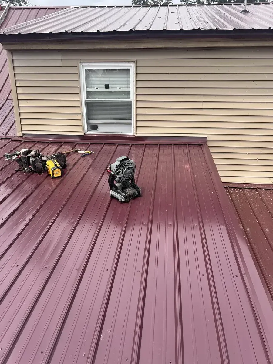 A pair of work boots and a small power tool sit on a maroon metal roof below a beige-sided house window.