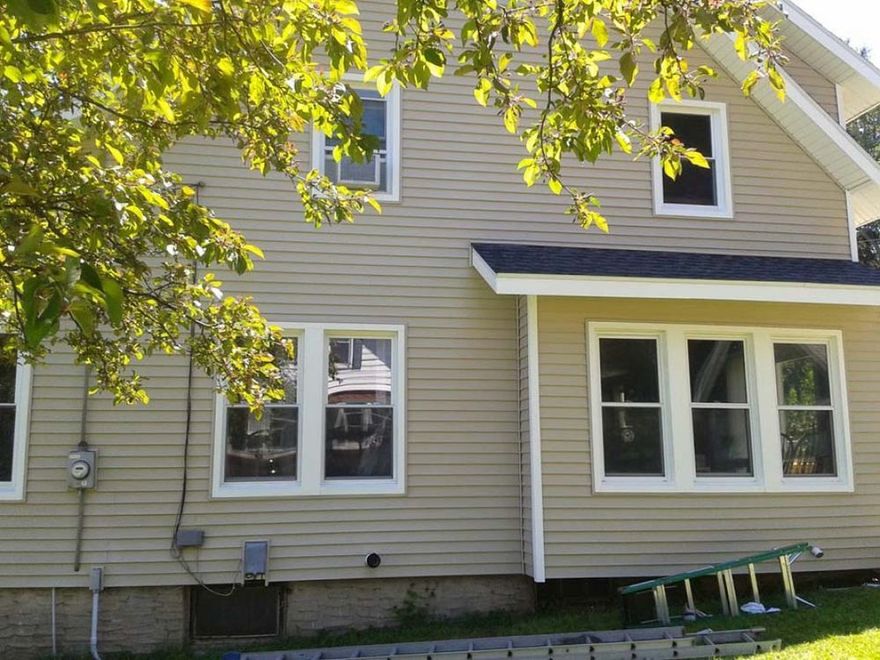 Tan vinyl-sided house exterior with several white-framed windows, a partially visible porch, and a tree branch in front.
