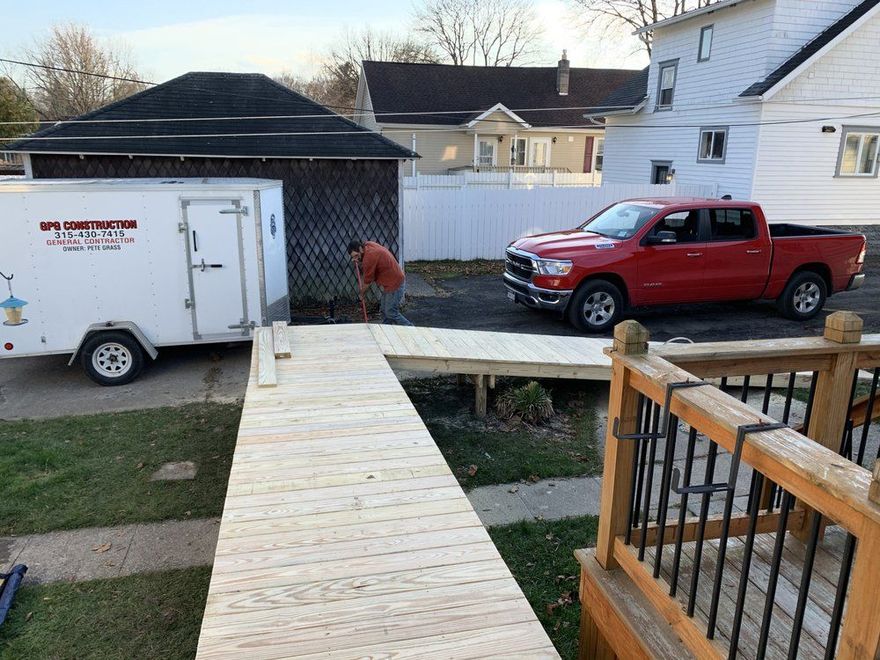 A worker builds a wooden wheelchair ramp leading to a deck, with a utility trailer and red truck parked nearby.