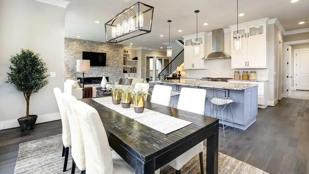 An open-concept room features a dark dining table with white chairs, a kitchen island, and a fireplace with a TV.