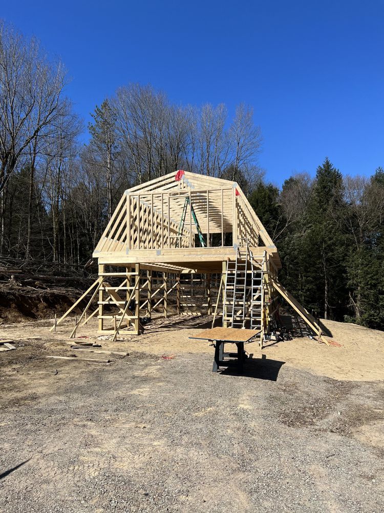 A wooden barn frame under construction on a gravel lot against a clear blue sky and a background of tall trees.